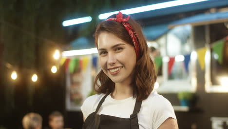 Close-up-of-Caucasian-beautiful-young-woman-in-apron-smiling-happily-to-camera-outdoors-with-little-food-truck-bar-on-background.-Portrait-of-pretty-female-bartender-or-vendor-at-fair-in-park.